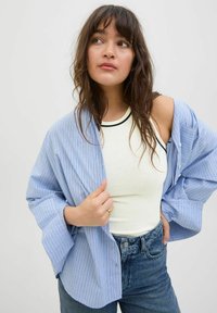Stribet blå skjorte med løse ærmer over en hvid tanktop. Blå denimjeans med klassisk knaplukning. Lys, afslappet tekstur.