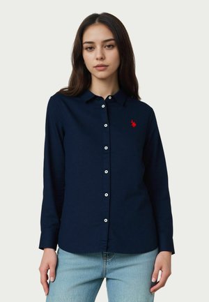 Giovane donna che indossa una camicia blu navy a maniche lunghe con bottoni e un logo ricamato rosso sul petto, abbinata a jeans azzurri.