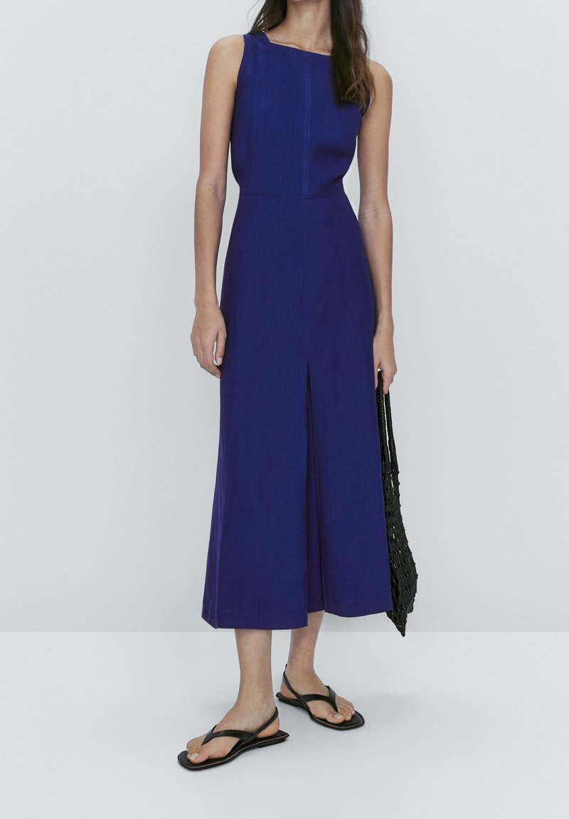 Femme portant une robe midi bleu foncé sans manches avec une fente à l'avant, tenant un sac cabas noir en filet, et portant des sandales plates noires.