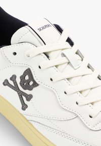 Zapatillas de cuero blanco con diseño de calavera y tibias en gris. Cuenta con cordones planos blancos, suela de goma y el logo "SCALPERS" en la lengüeta.
