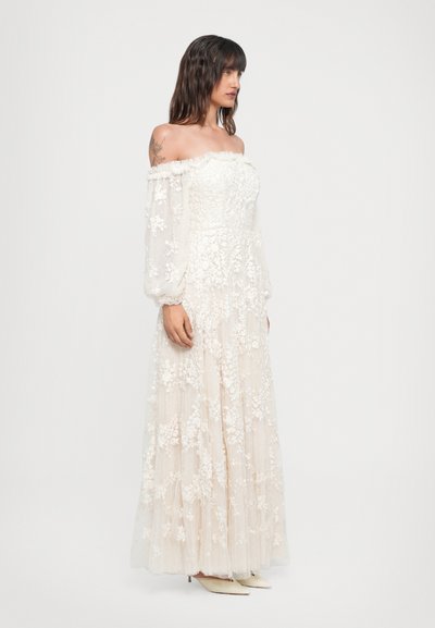 Vestido beige off-the-shoulder con intrincados bordados florales, tejido texturizado, mangas largas, falda en capas y tacones blancos puntiagudos.