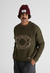 Knit sweater in dark green with a large beige circular pattern at the center. Features ribbed crew neck and frayed hem detail.