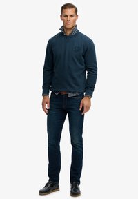 Granatowy sweatshirt o teksturowanym wykończeniu, z logo, noszony pod koszulą w kratę. Połączony z ciemnym dżinsami i czarnymi butami.