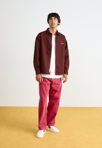 Μαρόν πουκαμίσα πάνω από ένα λευκό μπλουζάκι, συνδυασμένη με ροζ τζιν και λευκά αθλητικά παπούτσια, στέκεται σε κίτρινο πάτωμα.