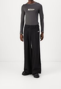 Osoba v šedém dlouhém rukávu s bílým logem a černých kalhotách s širokými nohavicemi s logem, stojící na bílém podlaze proti jednoduchému bílému pozadí.