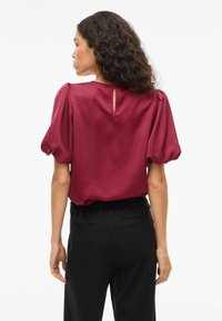 Blouse en satin bordeaux avec manches bouffantes et ouverture en trou de serrure dans le dos, associée à un pantalon noir avec poches latérales et taille élastique.