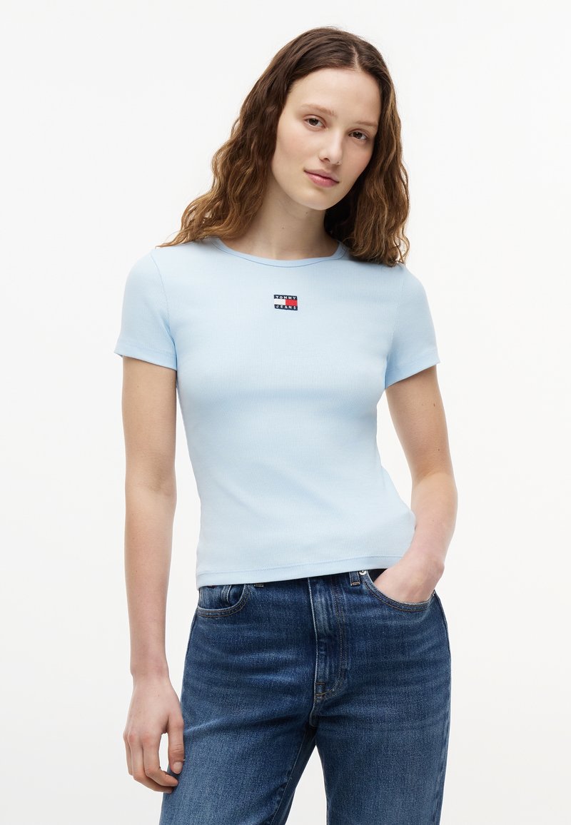 Jeune femme aux cheveux bruns ondulés portant une chemise ajustée à manches courtes bleu clair et un jean bleu, debout avec une main dans la poche.