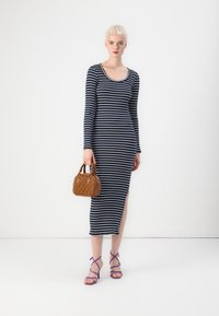 Ριγέ φόρεμα με μακρύ μανίκι σε ναυτικό μπλε και λευκό, που διαθέτει και μια πλευρική σχισμή. Το μοντέλο κρατά μια καφέ υφασματινή τσάντα και φορά μωβ σανδάλια με λουριά.