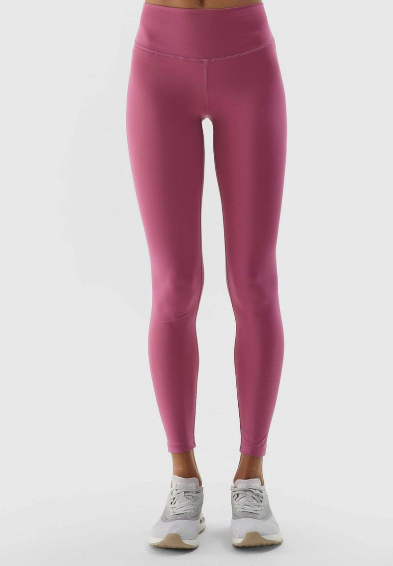 Fuchsia Leggings mit hohem Bund, aus dehnbarem Material. Getragen mit hellen Sportschuhen mit strukturiertem Sohlen.