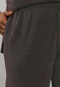 Hand rustend in de voorzak van donkere broek met multiculturele verticale strepen en een getailleerde pasvorm.