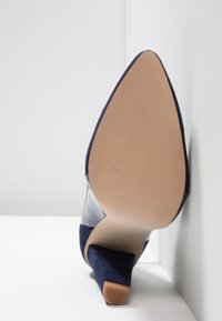 Chaussure à talons hauts bleu marine à bout pointu reposant sur le côté, montrant la semelle lisse et le talon bloc sur fond blanc.