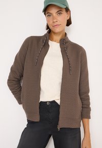 Giacca marrone con chiusura a zip in tessuto strutturato, colletto alto e coulisse, abbinata a una camicia crema e jeans neri. Il modello indossa un cappellino verde.
