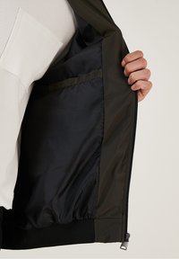 Tumma vihreä pommittaja-takki mustalla vuorilla, täysipitkä vetskari ja joustavat hihansuut. Oikealla puolella on sisätasku.