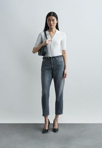 Γυναίκα που φοράει λευκό πλεκτό τοπ, ψηλόμεσα γκρίζα τζιν με κουμπιά, μαύρη ζώνη, μαύρες ψηλοτάκουνες γόβες και κρατά μαύρη τσάντα ώμου, στέκεται μπροστά σε απλό φόντο.