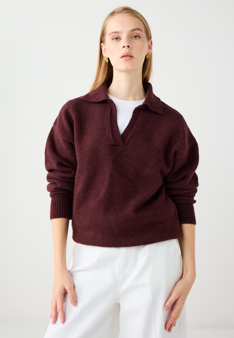 Burgundowy sweter dziany z kołnierzem, w fasonie V-neck i ściągaczami przy mankietach. Noszony na białej koszuli i zestawiony z białymi spodniami.