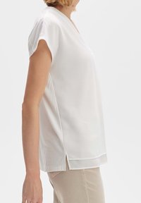 Blouse blanche à manches courtes avec un léger col en V, fabriquée en tissu léger et lisse. Comprend des fentes latérales à l'ourlet pour plus de confort.