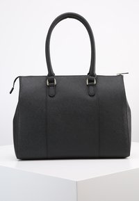 Czarna, strukturalna torebka na ramię z podwójnymi uchwytami, teksturowaną powierzchnią i zamknięciem na górze, umieszczona na białym piedestale wystawowym.