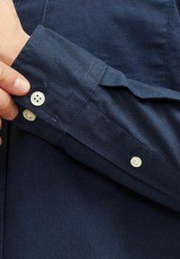 Puño de camisa azul marino con una tela texturada, que presenta dos botones blancos y un cuello en punta. Detalle de costura visible en el puño.