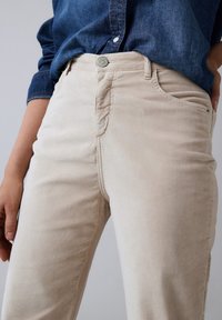 Pantalones de pana beige con corte recto, que cuentan con bolsillos frontales y cierre de botón. La tela tiene una textura suave y un brillo sutil.