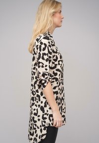 Leopardenmuster-Bluse mit beigem Hintergrund und schwarzen Flecken. Sie verfügt über eine lockere Passform, hochgerollte Ärmel und einen längeren Rücken-Saum.