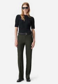 Zwart geribd gebreid topje, olijfgroene op maat gemaakte broek, zwarte riem en zwarte schoenen. Model draagt zwarte rechthoekige zonnebril.