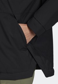 Main insérée dans la poche latérale zippée d'une veste noire avec une manche à revers, portée sur un pantalon vert olive.