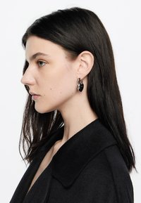 Joven mujer con cabello oscuro que lleva pendientes en forma de candado en forma de corazón negro y un abrigo negro, mirando hacia la izquierda sobre un fondo blanco.