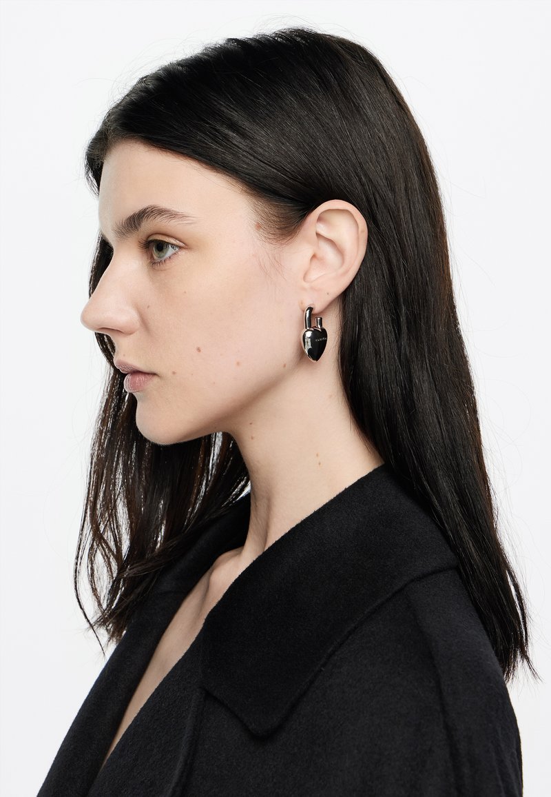 Joven mujer con cabello oscuro que lleva pendientes en forma de candado en forma de corazón negro y un abrigo negro, mirando hacia la izquierda sobre un fondo blanco.