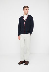 Σκούρο μπλέ κάρντιγκαν με V λαιμόκοψη, διακοσμημένο με κόκκινες και λευκές ρίγες κατά μήκος του γιακά και της γραμμής των κουμπιών, συνδυασμένο με ανοιχτόχρωμο παντελόνι.