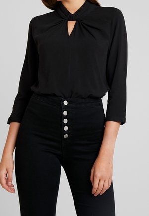 Femme portant un chemisier noir à encolure clé torsadée avec des manches trois-quarts et un pantalon noir taille haute à cinq boutons argentés.