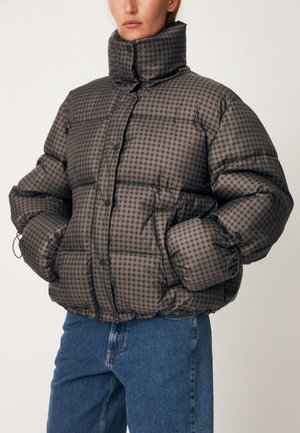 Femme portant une doudoune marron à carreaux avec un col montant, les mains dans les poches et un jean bleu sur un fond uni.