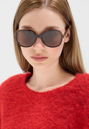 Giovane donna con capelli lisci castano chiaro indossa occhiali da sole scuri oversize e un maglione rosso peloso, guardando avanti su uno sfondo semplice.