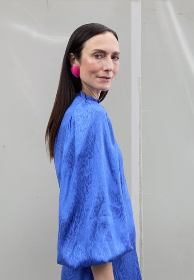 Blauwe blouse met print en opgepofte mouwen, voorzien van een hoge halslijn. Grote roze oorbellen in hartvorm zorgen voor kleurcontrast.