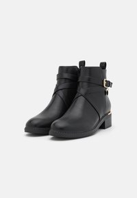 Bottines noires à la finition en cuir lisse, avec des sangles croisées, un panneau latéral texturé et un petit talon carré. Ornementation en métal doré.
