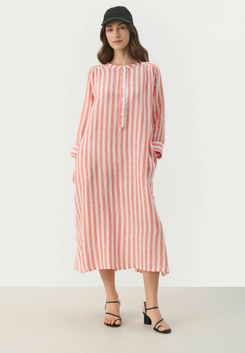 Gestreiftes Leinenkleid in Korall und Weiß mit langen Ärmeln, Knopfdetail und Seitenschlitzen, kombiniert mit schwarzen High Heels Sandalen und einer Kappe.