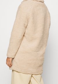Lysebeige faux fur jakke med en struktureret, fluffy overflade. Har en krave og knælangt design. Ingen synlige lukninger eller dekorationer.