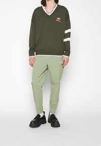 Osoba v olivově zeleném Nike svetr s V výstřihem a bílými pruhy, doplněná světle zelenými Nike kalhotami, černými robustními botami a bílými ponožkami.