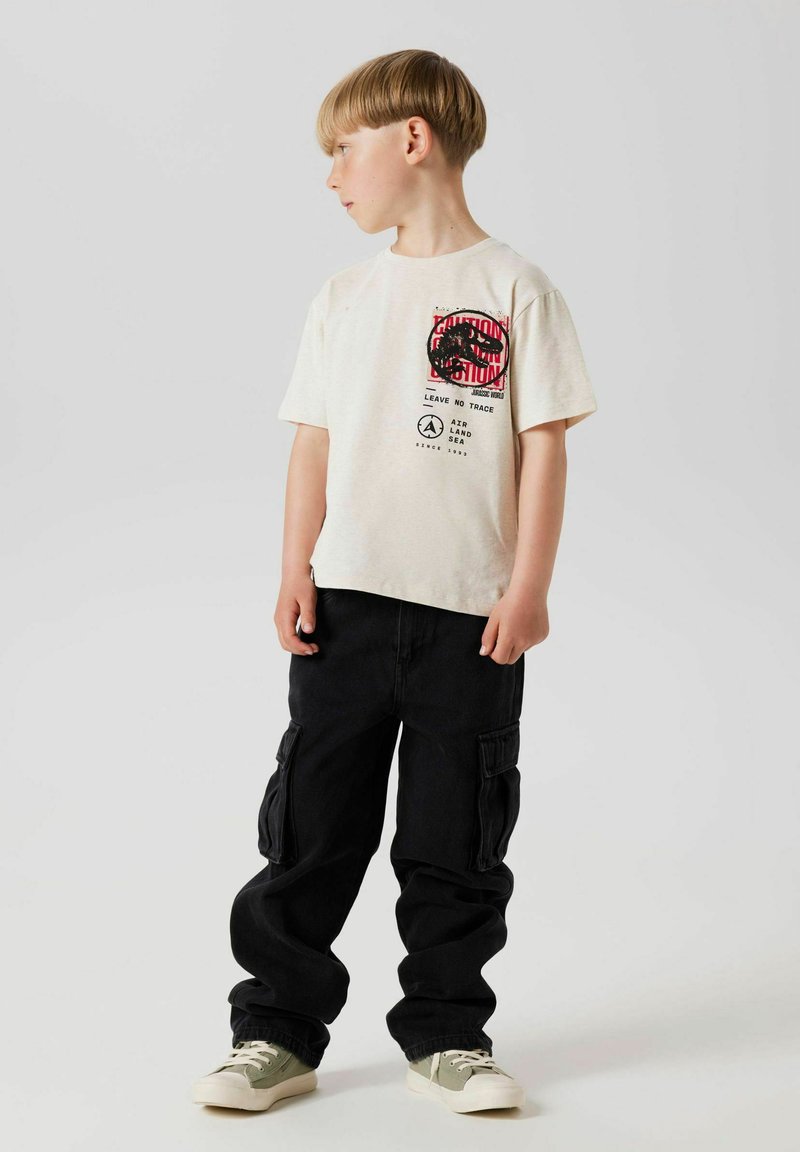 Camiseta gráfica de un beige claro con estampado en negro y rojo, combinada con pantalones cargo negros y zapatillas verdes. La camiseta tiene un corte relajado.