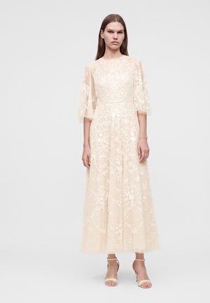 Vrouw die een lange, doorschijnende, crèmekleurige bloemenkant jurk draagt met ellebooglange mouwen en beige sandalen met hoge hakken op een effen achtergrond.