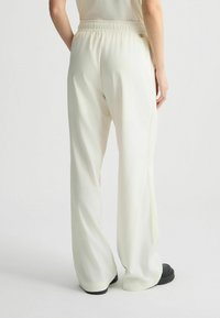 Creme broek met elastische tailleband en een relaxte fit, zachte textuur en een subtiele glans. Heeft zijnaad en een kleine metalen accent.