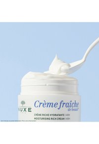 Vit burk med fuktighetskräm med en slät, krämig konsistens. En sked tar upp produkten. Etiketten har blå och gröna texter.