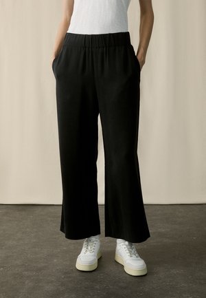 Personne portant un pantalon noir à jambes larges avec une taille élastique et des baskets blanches, debout sur un sol gris contre un mur beige.