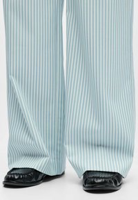 Helehallid triibulised laia lõikega püksid, valmistatud siledast kangast, kombineeritud mustade lahtiste kingadega, mille servadel on kogutud tekstuur.