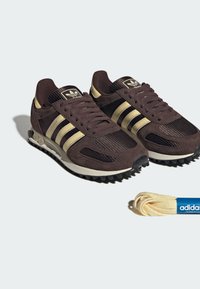 Baskets Adidas marron foncé avec des bandes beige et des lacets extra beige assortis placés à côté sur un fond clair.