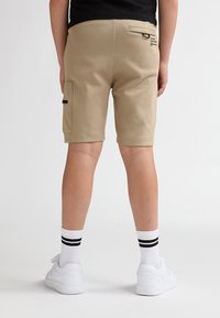 Beige shorts med sidelomme, elastisk talje og baglomme. Båret med hvide sneakers og stribede sokker, set bagfra.