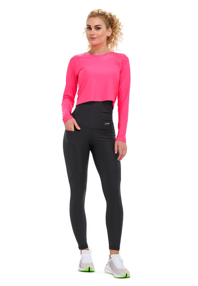 Top corto a maniche lunghe di un brillante colore rosa con leggings neri a vita alta dotati di tasche laterali. Tessuto texturizzato abbinato a scarpe da ginnastica di colore contrastante.