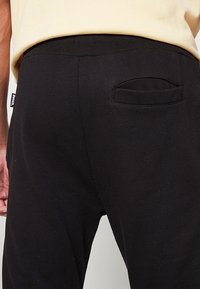 Pantalon de survêtement noir avec une taille élastique et une poche arrière ovale unique. Le tissu semble doux avec une légère texture.