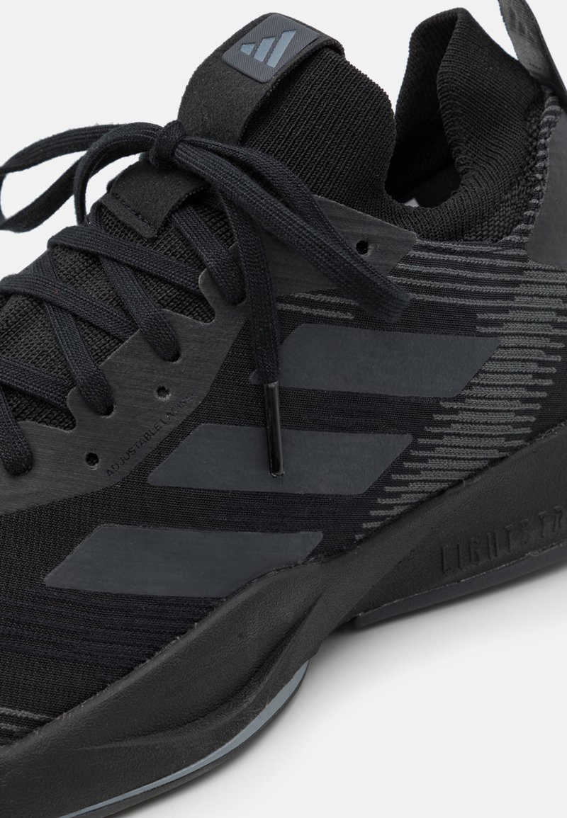 Zapatilla deportiva negra con parte superior texturizada, detalles en gris, un collar acolchado y cordones finos. Contiene una suela flexible para mayor soporte.