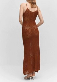 Femme de dos portant une robe maxi en crochet marron sans manches et des sandales à talons blancs, devant un fond uni.