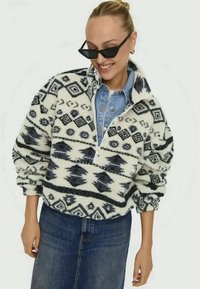 Pullover in pile con colletto a zip, caratterizzato da un motivo geometrico in bianco e nero. Tessuto morbido, vestibilità over size, abbinato a una camicia di denim.